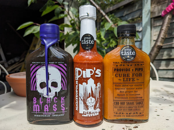 Three bottles of Pip's Hot Sauce arranged side by side on an outdoor table. From left to right: 'Black Mass' with a purple wax seal and gothic skull design, 'Pip's' hot sauce with a white wax seal and skeleton rider label, and 'Provide x Pip's Cure for Life' with a black cap and humorous label promoting the sauce's exaggerated benefits. All bottles feature 'Great Taste' award stickers and are set against a background of green foliage and a garden shed.