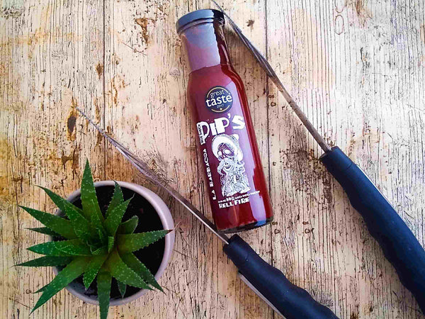 A bottle of "Pip's La Barbacoa Hellfire BBQ Sauce" lying on a rustic wooden surface next to a potted succulent and two large kitchen knives. The label features a skeleton graphic and the text "Great Taste 2017," highlighting the sauce’s smoky and fiery flavour.