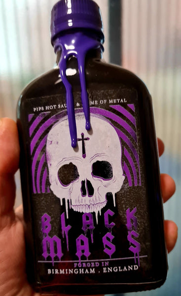 A close-up of a dark glass bottle with a purple wax seal dripping down the cap, featuring a skull design on the label with the text "Black Mass." The label also includes "Pip's Hot Sauce & Home of Metal" and "Forged in Birmingham, England."