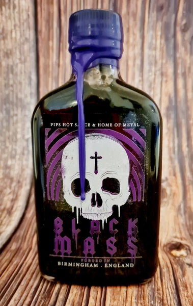 A close-up shot of a dark glass bottle with a striking label that features a white skull with a purple cross on its forehead. The bottle is sealed with purple wax, and the label reads "Black Mass," indicating it's a collaboration between Pip's Hot Sauce and Home of Metal. The background suggests a rustic setting, enhancing the bold and edgy vibe of the product.