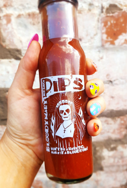 A hand holding a bottle of Pip’s Bloody Mary Hot Sauce against a brick wall background. The label features a skeleton wearing a nun's habit surrounded by flames, with the text "Don't be a heretic, make it a Bloody one!" The person’s nails are painted in vibrant colors, with each nail featuring a different playful design.