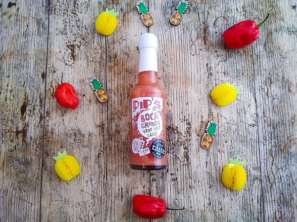 A bottle of Pip’s Boca Grande Very Hot Sauce placed on a rustic wooden surface. The bottle is surrounded by a circle of small yellow pineapple and red chili pepper decorations, creating a festive arrangement. The label on the bottle features the text "Pip's Boca Grande Very Hot Sauce" and a "Great Taste" award sticker.