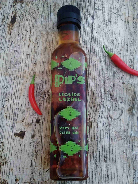 A bottle of Pip's Liquido Luzbel very hot chili oil resting on a rustic wooden surface, with a small red chili pepper lying beside it. The bottle's label is decorated with neon green designs and text, indicating the sauce's spicy nature.