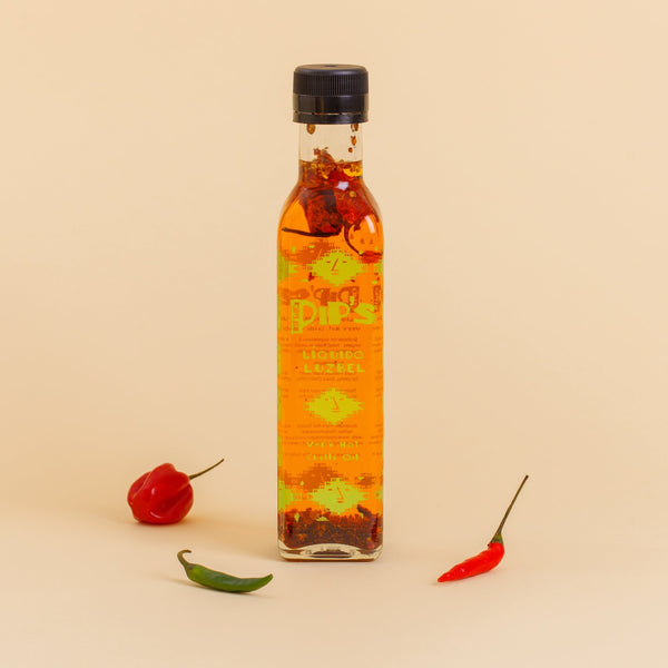 A bottle of Pip's Liquido Luzbel very hot chili oil standing upright against a neutral beige background. Two chili peppers, one green and one red, are placed beside the bottle, emphasizing the sauce's fiery spiciness. The oil inside the bottle shows visible chili flakes, enhancing the visual appeal of its heat.