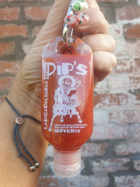 emergency hot sauce bottle of "pip's La Boca del Diablo Hot Sauce" being held in someone's hand with painted nails with chilli pepper designs, adding a playful touch. The bottle features a skeleton graphic and the text "There’s no smoke without fire. Home smoked and homemade. Super Hot." bright red hot sauce 