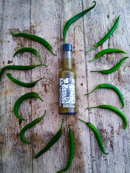 A bottle of "Pip's Fuego del Verde Hot Sauce" placed on a rustic wooden surface, surrounded by fresh green chilli peppers arranged in a circular pattern. The label features a striking design and the tagline "It's Green & Mean," emphasising the sauce's green, fiery characteristics.