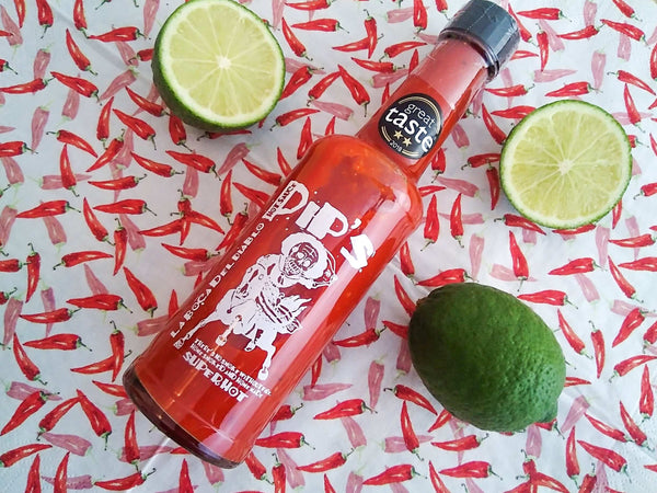 A bottle of Pip's "La Boca Del Diablo" super hot sauce, considered one of the best hot sauces, resting on a chilli-patterned background with lime accents. The bottle features a "Great Taste" 2018 award and the signature skeleton label, with the fiery red sauce inside.