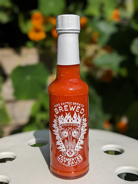 A bottle of Pip's X Attic Brew Co 'Brewed' fermented hot sauce with a distinctive white label featuring an intricate tribal design of a horned face and the words 'chilli peppers, garlic & onions.' The bottle is topped with a white wax seal and is set against a natural background with green foliage and orange flowers, creating a fresh and vibrant setting.