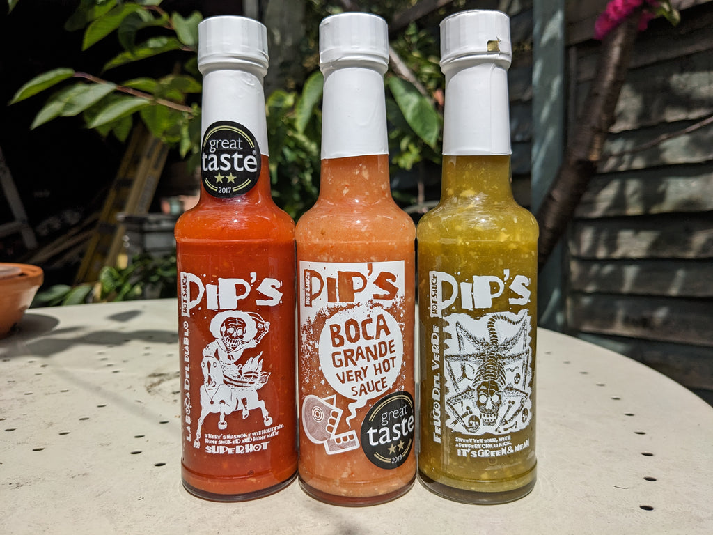 Three bottles of Pip's Hot Sauce arranged outdoors, each with distinct labels and vibrant colours. From left to right: "La Boca Del Diablo" in deep red, "Boca Grande" in bright orange, and "Fuego Del Verde" in a zesty green. Each bottle has won a "Great Taste" award, prominently displayed on the label.