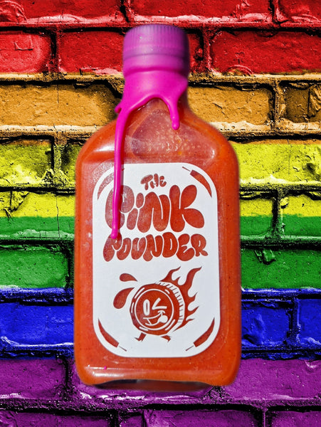 A bottle of 'The Pink Pounder' hot sauce with a pink wax seal dripping over the cap. The label features playful, retro-style text and a winking flame character. The bottle is set against a vibrant, rainbow-colored brick wall, adding a lively and colorful background to the image.