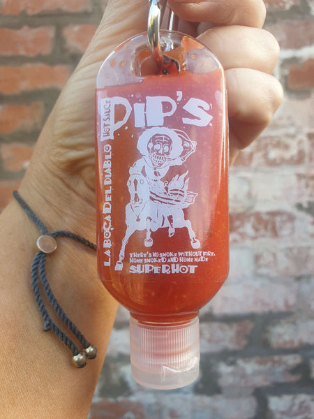 a small, travel-sized bottle of "Pip's La Boca del Diablo Hot Sauce" being held in someone's hand. The bottle features a playful skeleton graphic and the text "There’s no smoke without fire. Home smoked and homemade. Super Hot." The hot sauce is bright red, emphasising its spiciness. The background is a rustic, red brick wall.