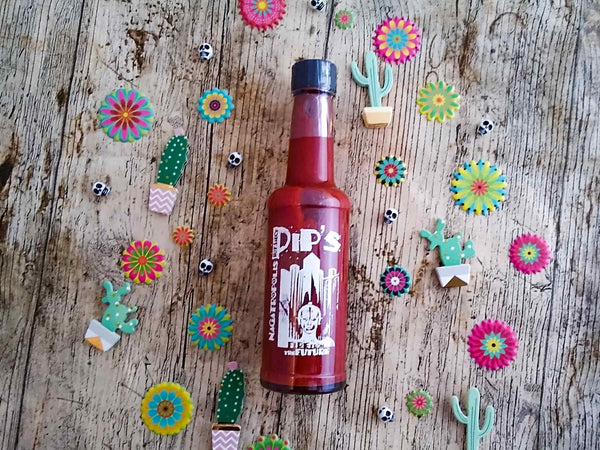 Pip's Nagatropolis Extra Hot Sauce bottle placed on a wooden surface, surrounded by colourful decorative cacti, skulls, and flowers, enhancing the hot and festive vibe of the product.