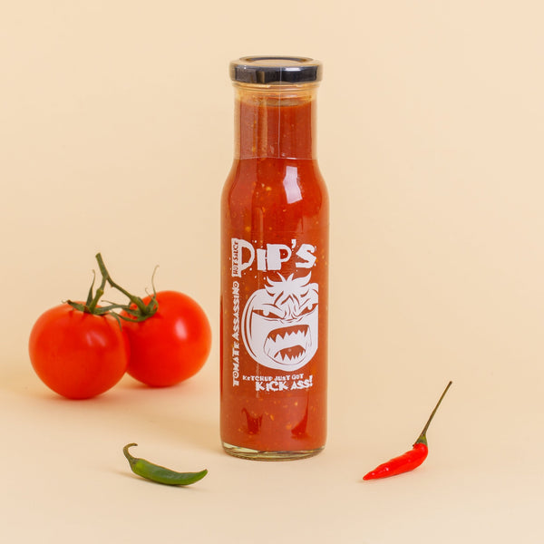 A bottle of Pip's Tomate Assassino spicy tomato ketchup with a vibrant red sauce, labelled with the slogan "Ketchup just got kick ass!" Two fresh tomatoes and chillies are positioned beside the bottle against a light backdrop.