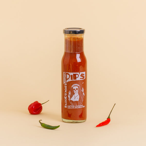 A bottle of Pip’s Bloody Mary Hot Sauce standing upright against a beige background. The label shows a skeleton in a nun's habit surrounded by flames, with the text "Don't be a heretic, make it a Bloody one!" The bottle is flanked by a red pepper on the left and a green and red chilli pepper on the right.