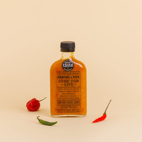  A bottle of Provide x Pip’s "Cure For Life" hot sauce, standing upright against a beige background. The label, which features a "Great Taste" award sticker, includes playful text such as "Gain third eye clarity" and "Improve sexual water," along with other whimsical claims. The bottle is flanked by a red chilli pepper on the left and a green and red chilli pepper on the right.