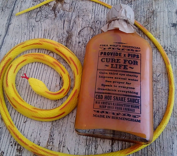 A bottle of Provide x Pip’s "Cure For Life" hot sauce lying on a rustic wooden surface. The label, featuring humorous claims like "Gain third eye clarity" and "Improve sexual water," is partially covered by a mock wax-sealed cap. A coiled yellow toy snake is placed beside the bottle, enhancing the quirky presentation.